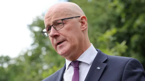 John Swinney is in the outdoors. There are trees with green leaves just out of focus behind him. Swinney is bald man and is wearing glasses, also a dark blue suit over a white shirt and purple tie. He has a pin badge of Scotland's saltire flag pinned to his lapel.