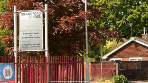 BBC The entrance and sign of the Blandford School. Trees can be seen around the buildings and the fence. It is a sunny day.