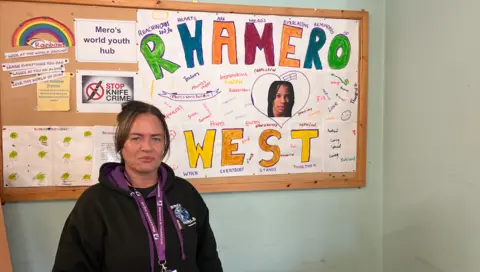 A woman stands in front of a board saying Rhamero West.