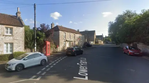 A Google Maps screenshot of Corston Post Office. The building is a pretty two-storey stone building, very similar to the others nearby. It is situated on a wide road next to a red phone box and a red post box, and has a small awning and a red sign. There are a couple of cars on the road. 