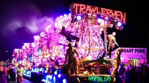 Illuminated carnival cart with purple, blue and red lights. There are people standing on it and a crowd is gathering around it.