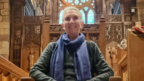 Stewart Bain Ann Cleeves, sits on a wooden chair inside what looks like a church. She is smiling and wearing a green jumper and purple scarf.