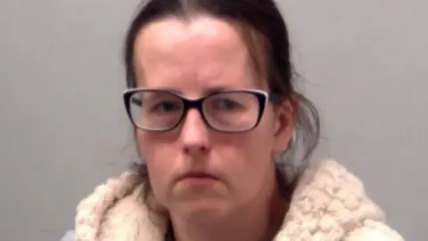 Essex Police A police custody image of Emma Barnett. She has dark brown hair pulled back from her head and black framed glasses. She is wearing a crime gilet over a grey T-shirt. 