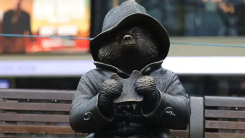 PA Media A statue of Paddington Bear, on a bench, eating a sandwich
