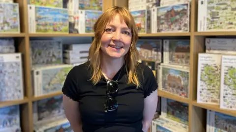A woman with long light coloured hair, is wearing a black top, with sunglasses clipped over the collar. She is standing in front of shelves full of jigsaw boxes