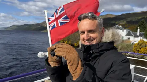 Man in a winter coat and gloves smiling while holding binoculars on a boat, with a red ensign flag flying behind him and a loch and hills in the background