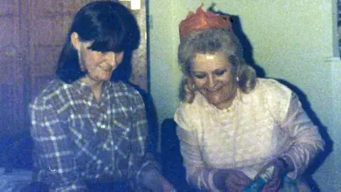 PA Media/family handout Christine and Patricia seated side by side indoors, positioned close together in front of a plain wall and a closed wooden door.
Christine is wearing a long‑sleeved, checked shirt and has dark hair that falls around the face. Patricia on the right is wearing a light‑coloured top and a paper party hat