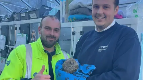South West Water George Cane and Joe Martin with Aquarius. Aquarius is sat on a blye blanket with black paw prints. Mr Cane is wearing a green hi-vis jacket and Mr Martin is wearing a blue jumper. 