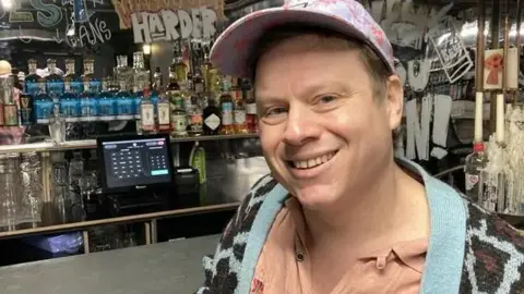 Mike is standing to the right of the picture, looking towards the camera smiling. He is wearing a blue and pink baseball cap, salmon pink colour top, and a blue and black cardigan. He is leaning against the bar in the club. 