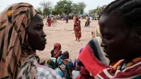 AFP via Getty Images Two Sudanese women, one wearing a headscarf, in the foreground. In the background, there are several other women and some are sitting on the ground. 
