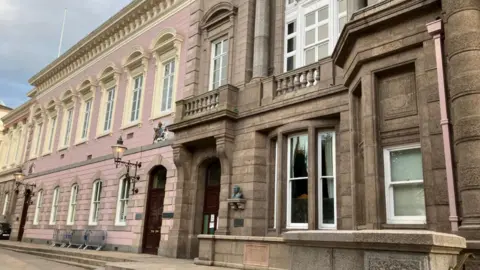 A picture of the outside of the Royal Court building in St Helier. There are two parts to the building, one is grey stone and the other is painted pink. 