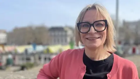 Neve Gordon-Farleigh/BBC Mel March standing at the top of Norwich Market at the memorial garden. She is looking directly at the camera and smiling and is wearing a black T-shirt and pink cardigan. Behind her is colourful stripey canopies. In the distance is Norwich castle.
