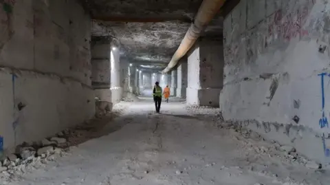 View along a large square horizontal mine shaft. Every surface is white Portland stone. Two men in hi-viz overalls and hardhats are walking away in the distance. The ceiling is about four times their height.