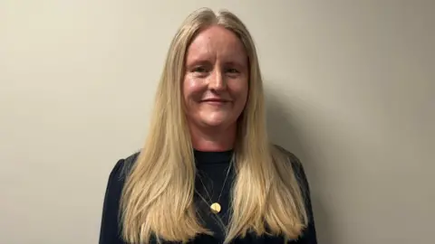 A woman with long blonde hair stands looking at the camera, she is wearing a black jumper and a gold necklace. 