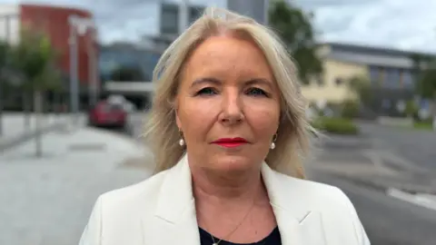 BBC Pat Cullen looks at the camera with a neutral expression on her face as she stands in the car park in front of South West Acute Hospital in Enniskillen but the background is blurred. She has shoulder-length blonde hair and is wearing red lipstick. She is wearing a white suit jacket coat and a black shirt and a pearl necklace and earring set.