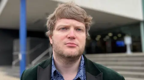 Tánaiste Custance has short brown hair. He wears a dark green velvet blazer over a blue paisley shirt and stands in front of a building