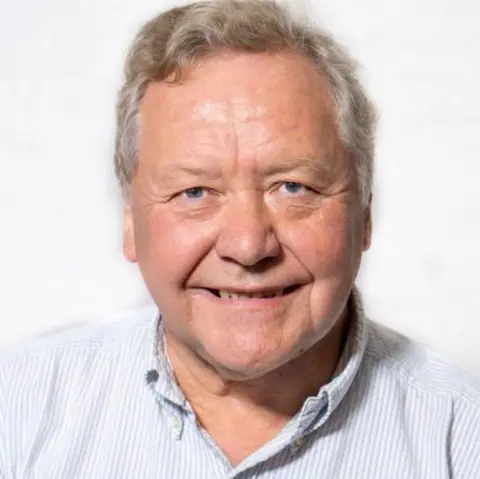 Staffordshire Moorlands Labour group A man in a striped blue-and-white shirt with an open collar smiles against a white backdrop. He has grey hair and blue eyes.
