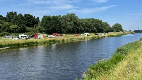 Andrew Turner/BBC Ambulance, police, fire and rescue appliances and cars and a caravan parked on the far bank of the river, with green banks either side and a row of trees behind on the far side. At the extreme right of the image is a sluice gate structure.