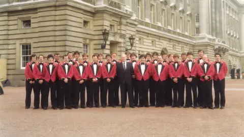 Foden's Band A group of musicians wearing red jackets, bow ties and black trousers, with one wearing a black suit and a tie, standing outside Buckingham Palace.