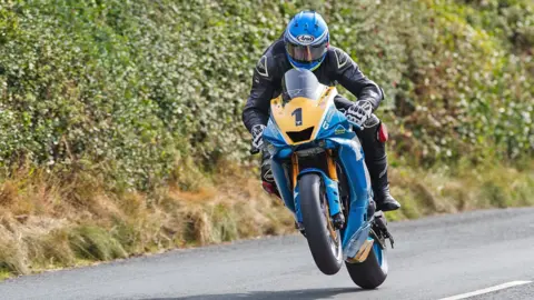 MANX GRAND PRIX Ingham riding in the Senior Manx Grand Prix in 2024. He is wearing black leathers and a blue helmet. The front wheel of the bike is lifting off the ground.