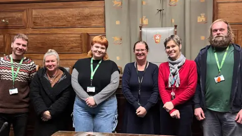 Maddy Jennings/BBC Six people standing in a row, smiling. There are two men and four women. They are all looking into the camera and have their hands together. In the background, there is a wooden wall and a grey curtain. 