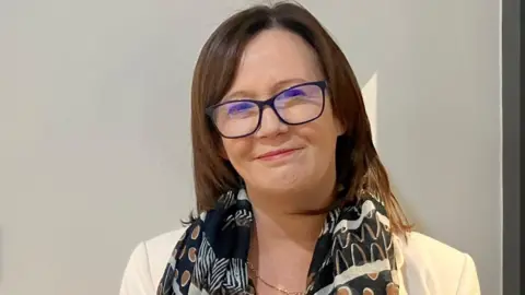 Eilis McGuinness  stands im front of a white wall. She has shoulder length dark hair and is wearing glasses, a scarf and white jacket