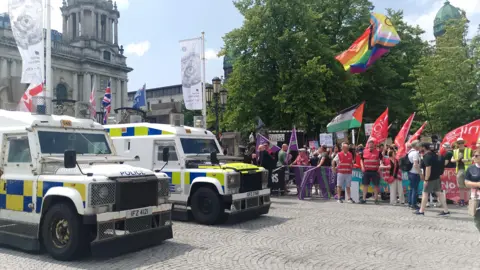 Police vehicles and protesters lined up outside Belfast City Hall during an anti-racism protest.  Two police Land Rovers are visible in the foreground.  The protesters are waving flags including red Nipsa flags. 