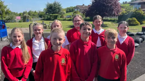 Nine children in red jumpers and white polo shirts standing on a playground with trees and grass in the background.