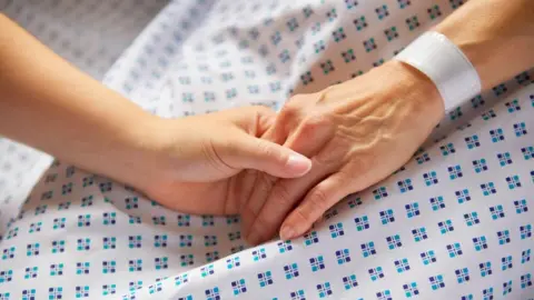 A hand holds the hand of a person in a hospital gown with a hospital wristband on
