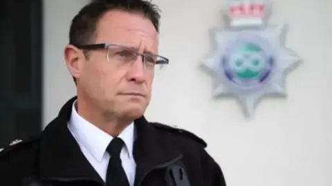 BBC Mr Noble is in uniform and is stood outside force headquarters being interviewed. He is wearing glass and a jacket.