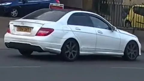 A CCTV still of a white Mercedes being driven along a road.