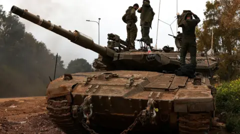 Israeli soldiers stand on top of a tank in northern Israel, near the Israel-Lebanon border. the tank is an imposing presence, taking up most of the frame.