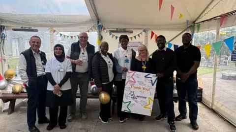 A group of people stand together inside a decorated tent, holding a “Thank You Volunteers Week” sign with bunting and balloons around them.
