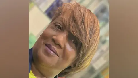 Close-up portrait of a person with short, side-swept light brown hair, looking toward the camera. The image is slightly tilted and framed tightly around the face and shoulders. The background is softly blurred, with indistinct indoor features visible behind the subject.