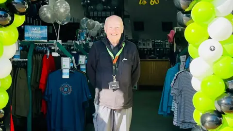 Doug smiles at the camera, standing underneath a balloon arch with rails fo clothing behind him. He wears a branded fleece and lanyards