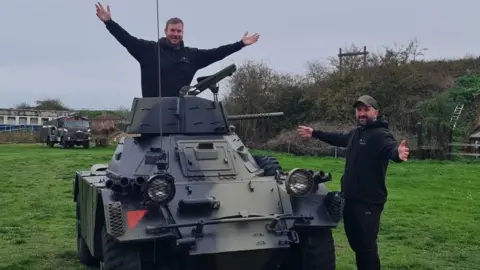 Nick Taylor A man stood, wearing a black hoodie, with his arms out on a Ferret armoured car. Another man is stood next to the vehicle with his arms out, wearing a black hoodie and a black cap. The vehicle is parked on grass.