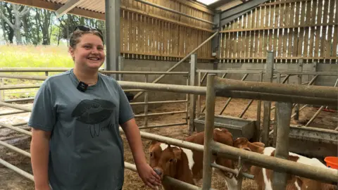 A teenage girl in a blue T-shirt with sunglasses on top of her head, standing in barn next to a pen with some brown and white dairy cows.