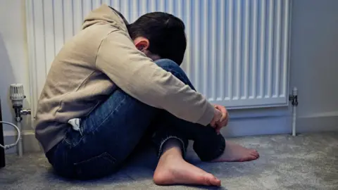 A child in jeans with a beige hooded top and bare feet has his head buried between his knees in front of a radiator