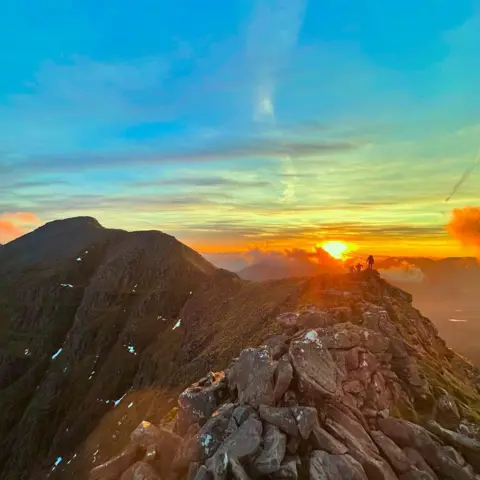 Daniel White A blue and orange sky above a rocky mountain. The silhouettes of three people are visible in the distance.