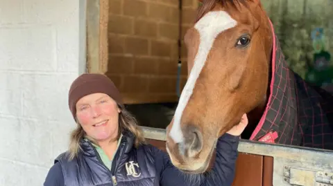 A picture of a woman stood outside a stable with her hand on a horse's chin. The brown horse is inside the stable and is wearing a dry rug.