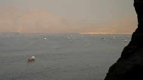 The Strait of Hormuz is shown from a dsitance w several tankers and freight boats. There are mountains in the foreground and in the distance.
