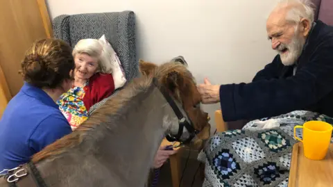 A group of three people are sat with a brown coloured miniature horse in the centre of the photo.  The horse is wearing a bridle. To the right there is an elderly man who is stroking the horse.  He is sat in a care home chair with a crochet blanket across his knees.  To the left of the picture his wife is sat in another care home chair.  She is talking to the horse's owner. The horse's owner has her back to the camera.  She has brown hair and is wearing a blue T-shirt.