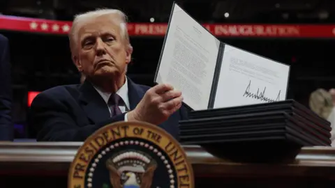 Reuters President Trump dressed in a dark suit with a dark tie and white shirt holds up a folder containing an executive order he has signed. He is sat a a table with the presidential seal in the foreground