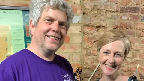 Ben Heaney, holding a violin, with Jessica Mogridge, holding an oboe, standing in front of a brick wall, Ben is wearing a purple T-shirt, has short grey hair and Jessica has short fair hair. They are both smiling.  