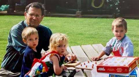 Caleb Tutt An older gentleman in a green shirt looks at the camera, along with three children. They are sat around a wooden picnic bench on a green. There are games on the bench. All three children also look at the camera.