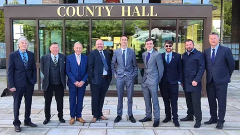 Great Yarmouth First Nine men, wearing a mixture of blue and grey suits, are standing outside a building marked COUNTY HALL