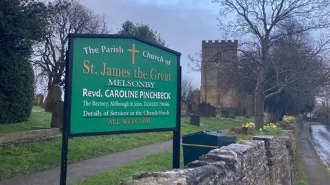 A rural churchyard scene on an overcast day. In the foreground, there is a large green sign for “The Parish Church of St James the Great, Melsonby.” The sign has gold and white lettering, including a gold cross at the top.