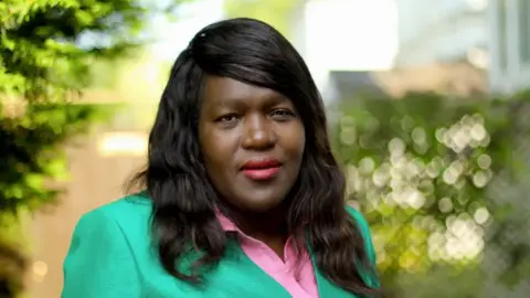 Florence Nyasamo A woman with black hair looks at the camera. She is wearing a green blazer with a pink shirt beneath it. There are hedges behind her.