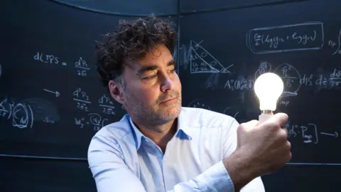 Martin Small Henry Snaith is wearing a light blue shirt and has short brown hair and brown stubble. He is holding a lit up lightbulb and behind him are chalkboards with mathematical formulas written on them