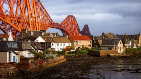 Getty Images The Forth Rail Bridge stretching behind a row of white and brown houses at sunset.